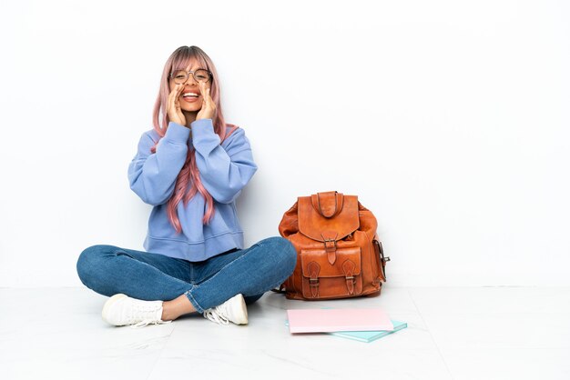 Giovane studentessa di razza mista con i capelli rosa seduta su un pavimento isolato su sfondo bianco gridando e annunciando qualcosa