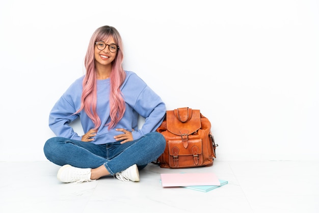 Giovane studentessa di razza mista con i capelli rosa seduta su un pavimento isolato su sfondo bianco in posa con le braccia all'anca e sorridente