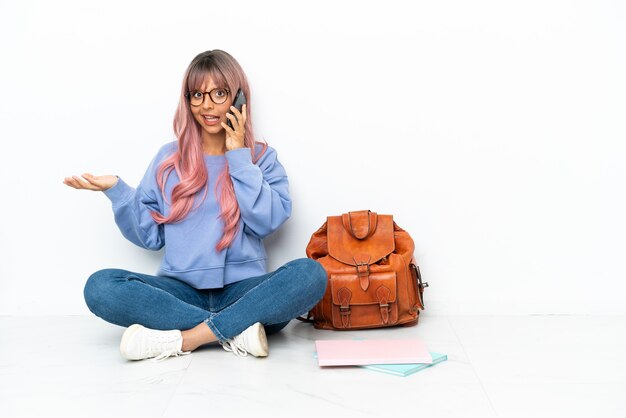 Giovane studentessa di razza mista con i capelli rosa seduta su un pavimento isolato su sfondo bianco mantenendo una conversazione con il telefono cellulare con qualcuno