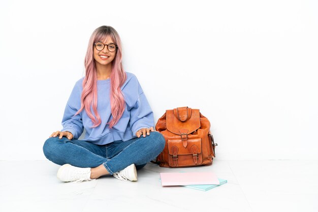 Giovane studentessa di razza mista con i capelli rosa seduta sul pavimento isolato su sfondo bianco ridendo