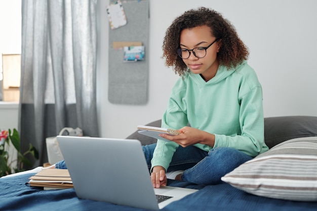 Giovane studentessa di razza mista in abbigliamento casual seduta sul letto davanti al laptop, leggendo note in quaderno e navigando in rete a casa
