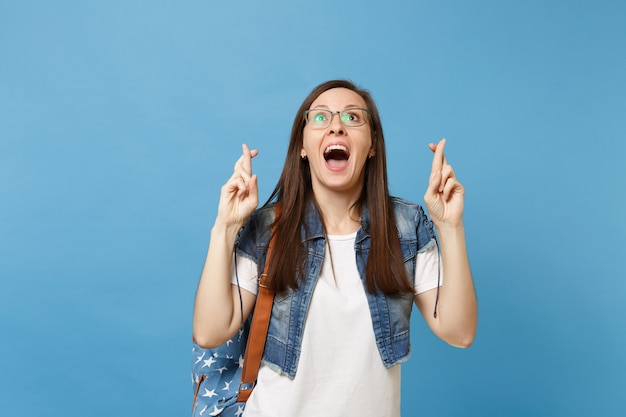 Giovane studentessa eccitata con gli occhiali con lo zaino in attesa di un momento speciale, tenendo le dita incrociate, alzando lo sguardo isolato su sfondo blu. Istruzione nel concetto di college universitario di liceo.