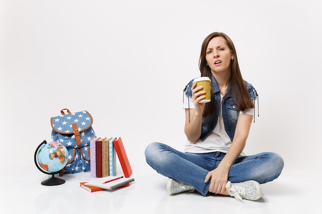 Giovane studentessa esaurita stanca che tiene tazza di carta con caffè o tè seduto vicino al globo, zaino, libri scolastici isolati