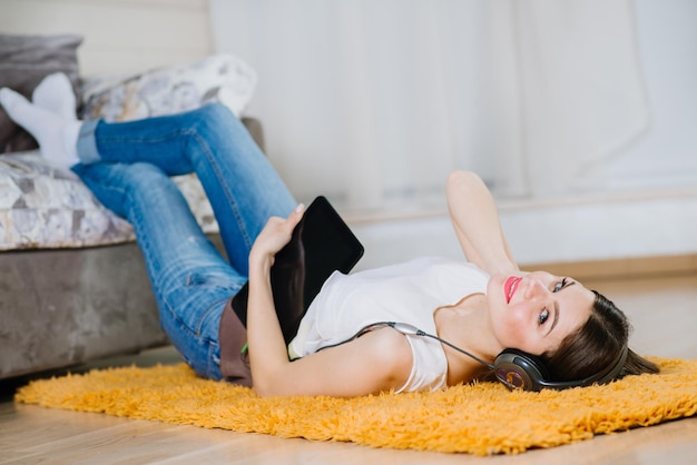 Giovane studentessa felice sdraiata sul pavimento e ascoltando musica dal tablet al chiuso a casa