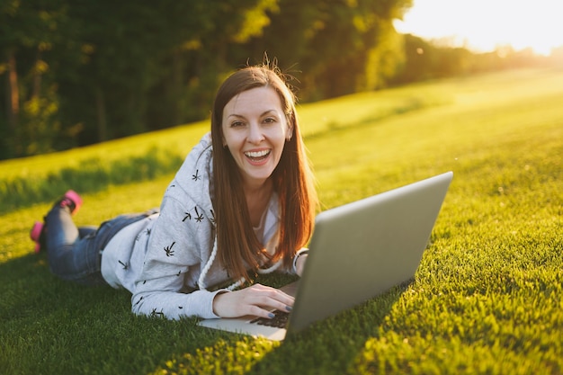 Giovane studentessa intelligente in abiti casual. Donna sdraiata sul terreno erboso, lavorando su computer pc portatile nel parco cittadino sul prato verde del sole dell'erba all'aperto. Ufficio Mobile. Concetto di business freelance.