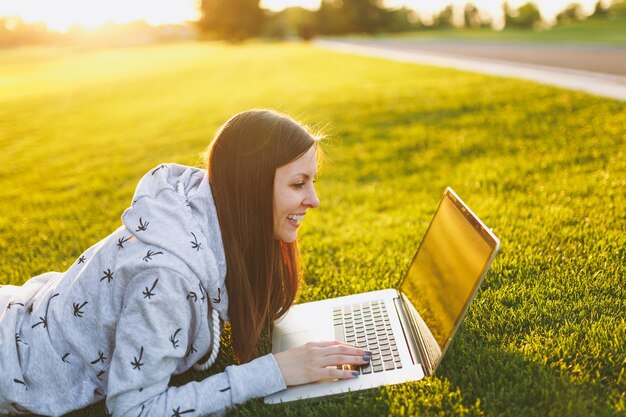 Giovane studentessa intelligente in abiti casual. Donna sdraiata sul terreno erboso, lavorando su computer pc portatile nel parco cittadino sul prato verde del sole dell'erba all'aperto. Ufficio Mobile. Concetto di business freelance.