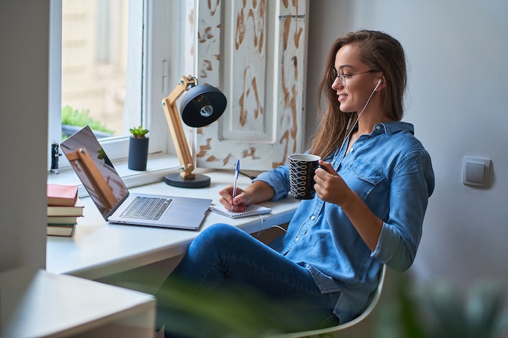 Foto giovane studentessa intelligente sorridente casuale in cuffie soddisfatta dell'apprendimento della lingua straniera. femmina che prende appunti al taccuino durante la visione di corsi video webinar. formazione online