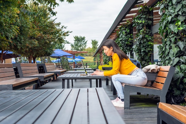 Giovane studentessa latina che indossa un maglione giallo usando il suo laptop seduto a un tavolo fuori