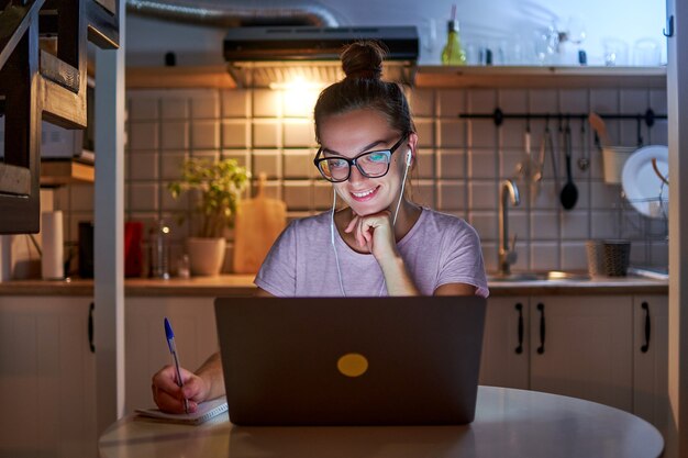 Giovane studentessa maniaco del lavoro in cuffia che si prepara per l'esame, prende appunti sul taccuino durante la visione del webinar e apprende una lezione online sul portatile a tarda sera