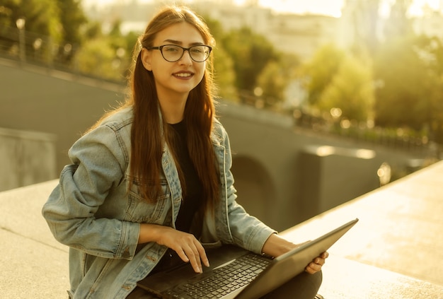 Giovane studentessa moderna in una giacca di jeans seduta con il computer portatile. Insegnamento a distanza. Concetto di gioventù moderna.