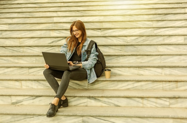 Giovane studentessa moderna in una giacca di jeans seduta sulle scale con il computer portatile. Insegnamento a distanza. Concetto di gioventù moderna.