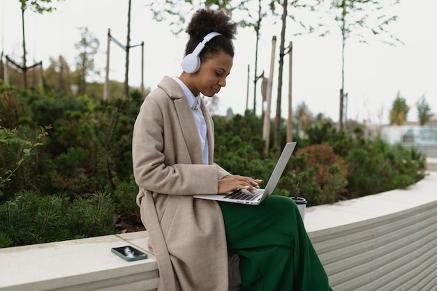 Giovane studentessa nera che si prepara per un esame lavorando su un laptop online in cuffia in una città