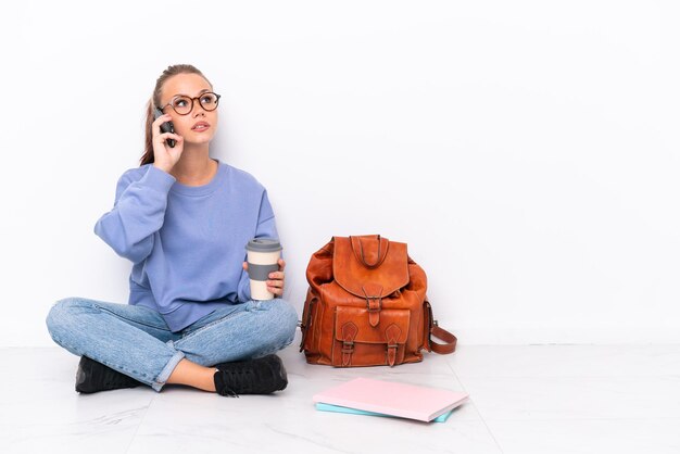 Giovane studentessa seduta su un pavimento isolato su sfondo bianco in possesso di caffè da portare via e un cellulare