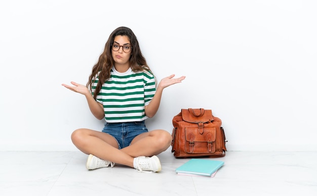 Giovane studentessa seduta sul pavimento isolata su sfondo bianco avendo dubbi mentre alza le mani