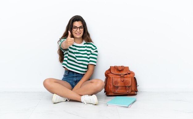 Giovane studentessa seduta sul pavimento isolata su sfondo bianco con il pollice in alto perché è successo qualcosa di buono