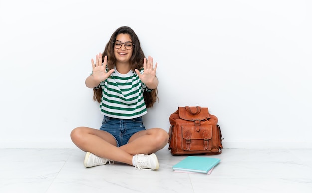 Giovane studentessa seduta sul pavimento isolata su sfondo bianco contando dieci con le dita