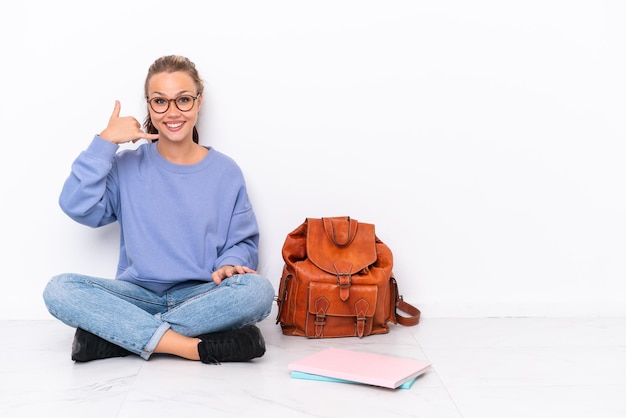 Giovane studentessa seduta sul pavimento isolato su sfondo bianco che fa il gesto del telefono Chiamami indietro segno