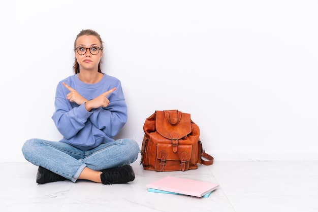 Giovane studentessa seduta sul pavimento isolato su sfondo bianco che indica i laterali che hanno dubbi