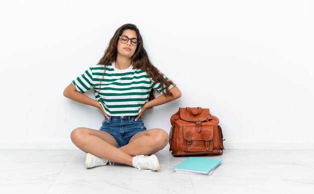 Giovane studentessa seduta sul pavimento isolato su sfondo bianco che soffre di mal di schiena per aver fatto uno sforzo