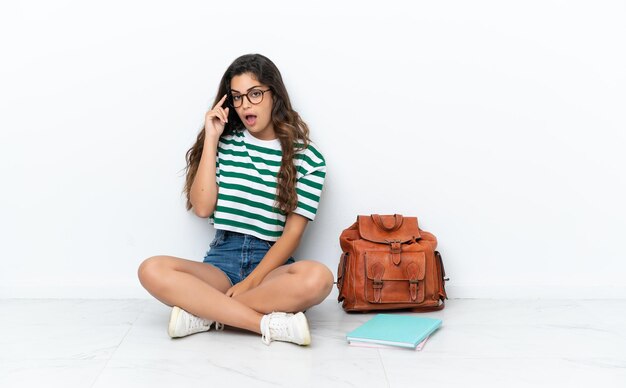 Giovane studentessa seduta uno sul pavimento isolato su sfondo bianco con gli occhiali e sorpreso