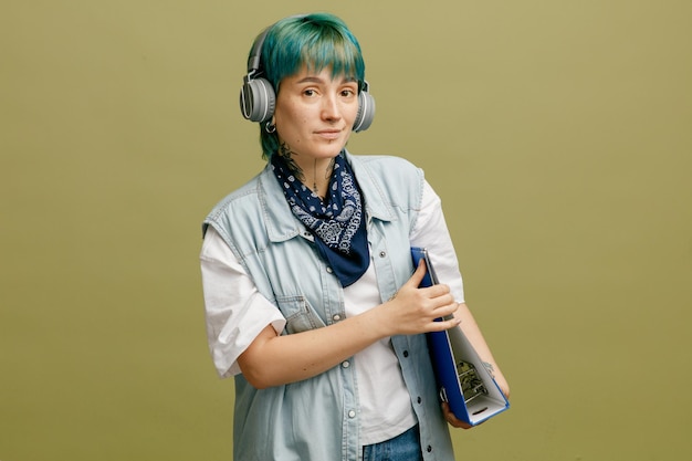 Giovane studentessa sicura che indossa le cuffie e la bandana sul collo che tiene la cartella con entrambe le mani guardando la fotocamera isolata su sfondo verde oliva