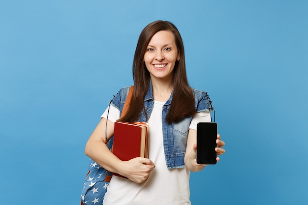 Giovane studentessa sorridente in vestiti di denim con zaino che tiene libri di scuola, telefono cellulare con schermo vuoto nero vuoto isolato su sfondo blu. Istruzione al college universitario di scuola superiore.