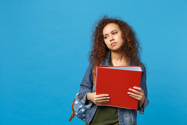 Giovane studentessa teenager della ragazza afroamericana in cartella della tenuta dello zaino dei vestiti del denim isolata sulla parete blu