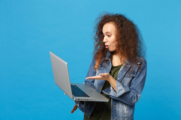 Giovane studentessa teenager della ragazza afroamericana in vestiti del denim, lavoro dello zaino sul pc isolato sulla parete blu