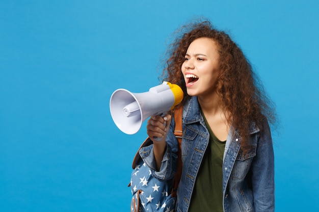 Giovane studentessa teenager della ragazza afroamericana in vestiti del denim, megafono della tenuta dello zaino isolato sulla parete blu