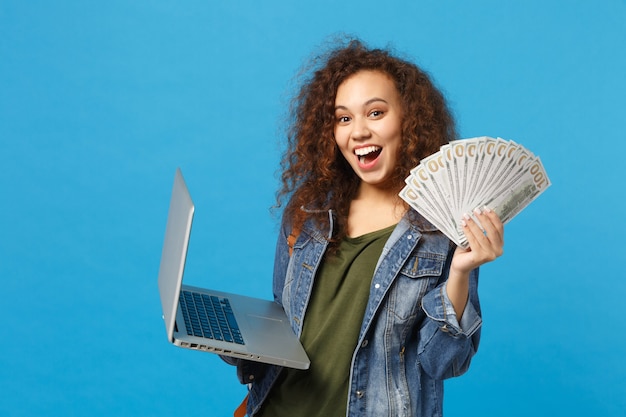 Giovane studentessa teenager della ragazza afroamericana in vestiti del denim, pc della tenuta dello zaino, fan di denaro contante isolato sulla parete blu