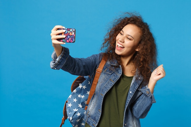 Giovane studentessa teenager della ragazza afroamericana in vestiti del denim, telefono della tenuta dello zaino isolato sulla parete blu