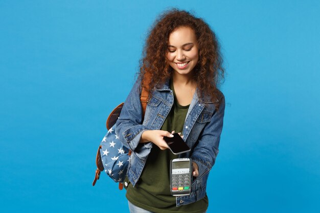 Giovane studentessa teenager della ragazza afroamericana in vestiti del denim, terminale della tenuta dello zaino isolato sulla parete blu