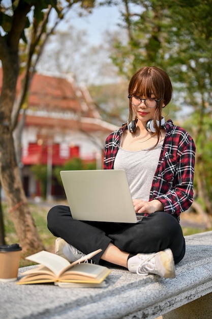 Giovane studentessa universitaria asiatica focalizzata che usa il computer portatile su una panchina nel parco del campus