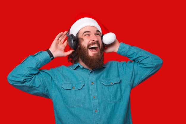 Giovane stupito con la barba che ascolta musica in cuffia e indossa il cappello di babbo natale