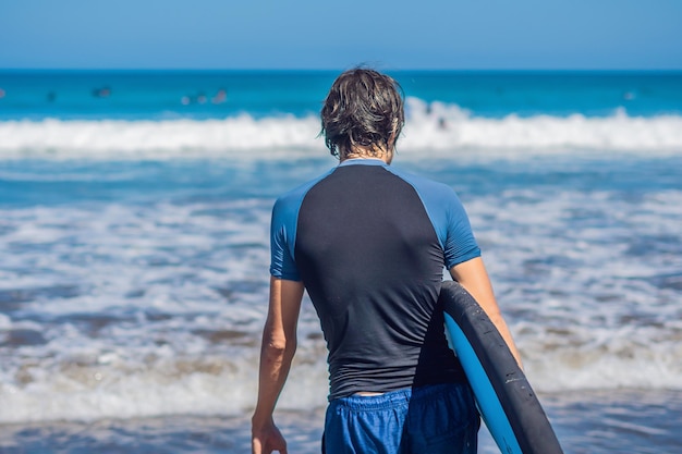 Giovane surfista sportivo bello che posa con la sua tavola da surf sotto il braccio nella sua muta su una spiaggia tropicale sabbiosa