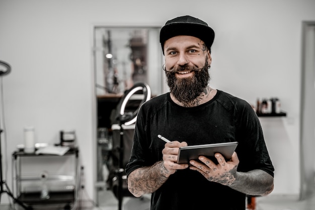Giovane tatuatore maschio con la barba che tiene matita e schizzo guardando positivo e felice in piedi e sorridente in officina.