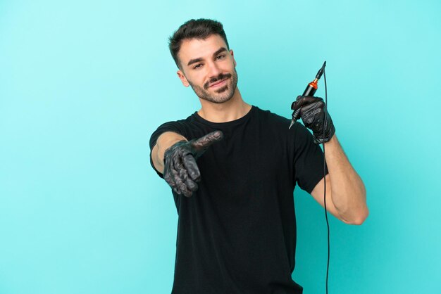 Giovane tatuatore uomo isolato su sfondo blu che stringe la mano per la chiusura di un buon affare