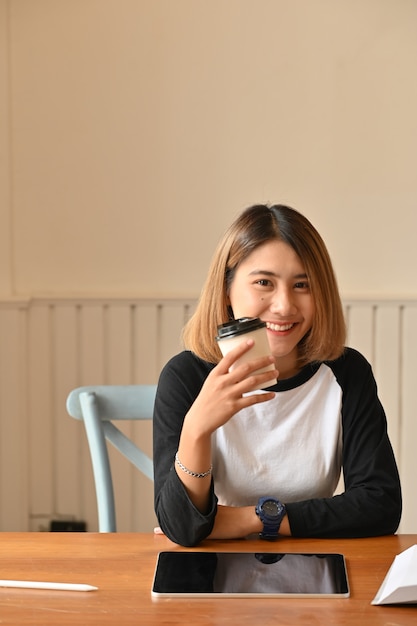 Giovane tazza di caffè attraente della tenuta della donna ed esaminare macchina fotografica sulla tavola creativa.