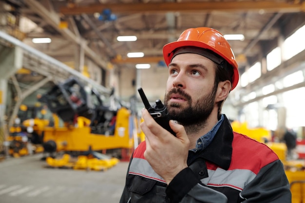 Giovane tecnico barbuto o operaio di fabbrica che parla nel walkie-talkie