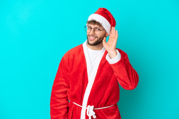 Giovane travestito da Babbo Natale isolato su sfondo blu che ascolta qualcosa mettendo la mano sull'orecchio