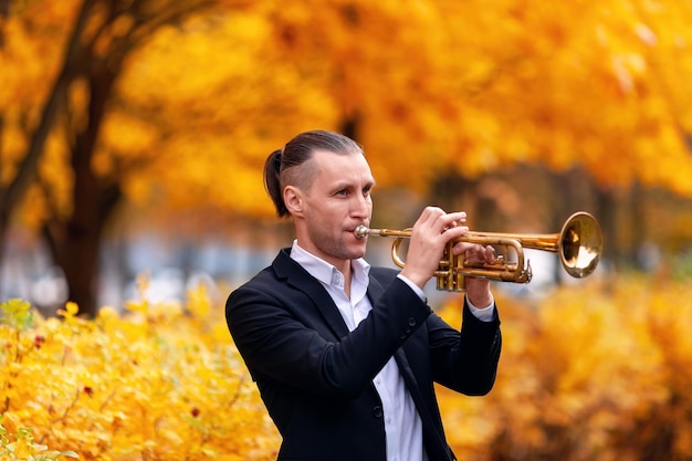 Giovane trombettista bello in abiti formali che suona il suo strumento musicale nel parco autunnale