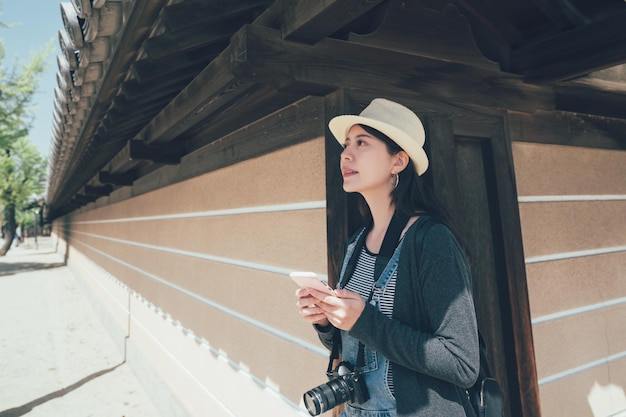 giovane turista asiatico che cammina attraverso il basso tetto di legno del muro nell'uscita del tempio di shitennoji. bella signora viaggiatore con cappello di paglia che tiene telefono cellulare e fotocamera lasciando lo shintoismo per trovare la direzione della mappa online