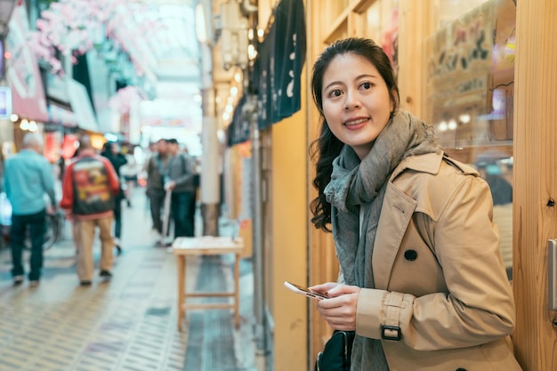 Giovane turista asiatico in cappotto che naviga nel sito web di Internet sul telefono cellulare e guarda da parte sulla strada nel mercato locale. ragazza appoggiata al muro del ristorante izakaya in attesa di un amico sorridente.