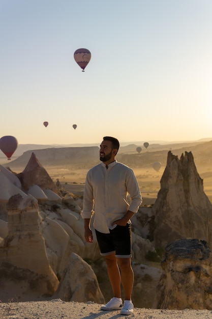 Giovane turista barbuto in primo piano con vista sulla valle dell'amore e mongolfiere che volano all'alba in Cappadocia, Turchia