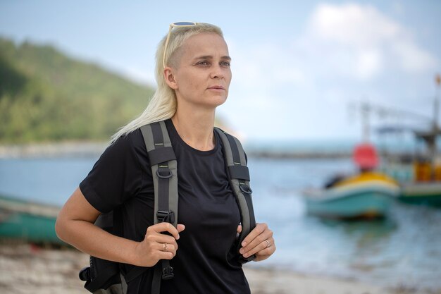 Giovane turista di una ragazza bionda con lo zaino sulla spiaggia con piacere guardando un bellissimo paesaggio marino, da vicino