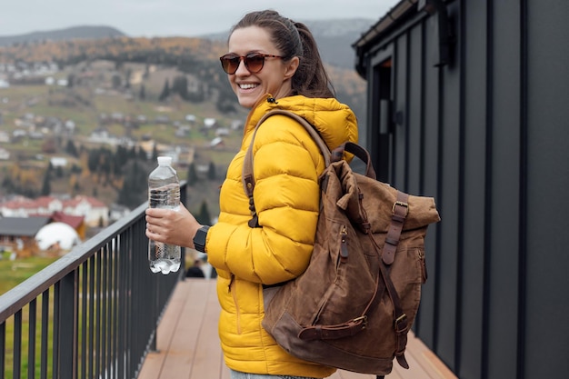 Giovane turista femminile in piumino giallo beve acqua fresca sullo sfondo della natura e delle montagne