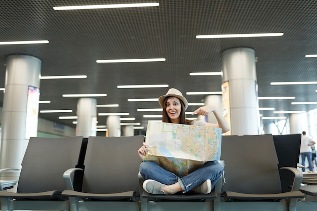 Giovane turista viaggiatrice con le gambe incrociate che punta il dito indice sulla mappa cartacea, in attesa nella hall dell'aeroporto internazionale international