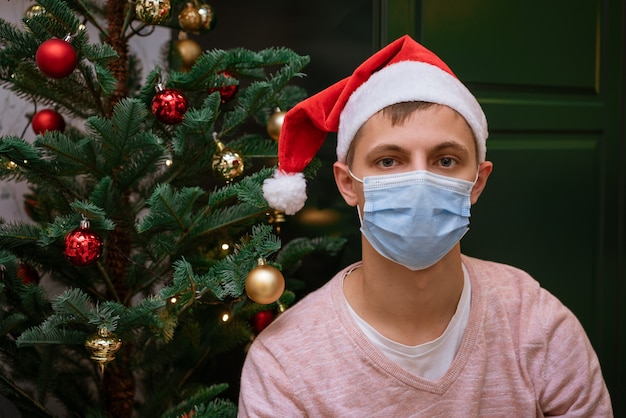 Giovane uomo a casa vicino a un albero di Natale che indossa una maschera e un cappello di Babbo Natale guarda la telecamera