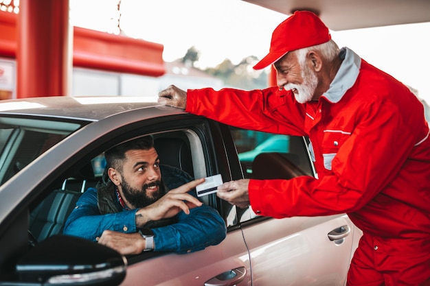 Giovane uomo adulto bello insieme al lavoratore anziano in piedi sulla stazione di servizio e fare rifornimento di auto e pagare con carta di credito.