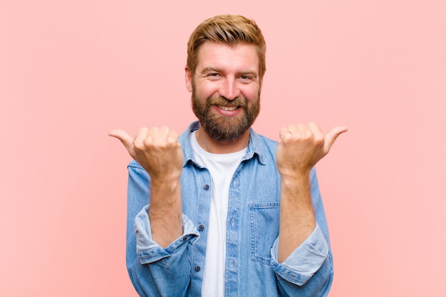 Giovane uomo adulto biondo che sorride allegro e che sembra felice, sentendosi spensierato e positivo con entrambi i pollici in su